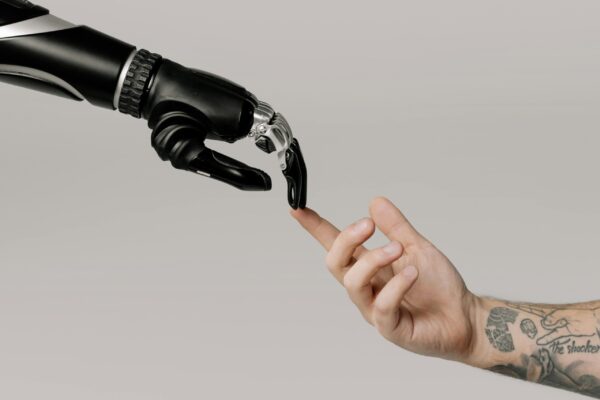 A robotic hand and a tattooed human hand reach out and touch fingertips against a plain light background, symbolizing human-robot connection.